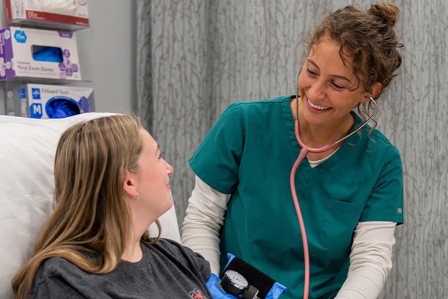 Nursing assistant student in teal scrubs smiling while taking a patient's blood pressure in a clinical lab